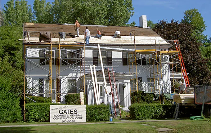 Roofers tearing off an old roof on a white colonial home, with a Gates Roofing project sign on the lawn