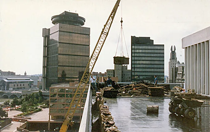 Crane hoisting debris from a downtown rooftop tear-off with city towers in the background