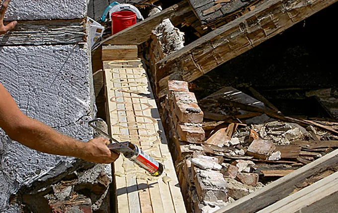 Close-up of a carpenter applying adhesive to a new rafter beside old brick and plaster