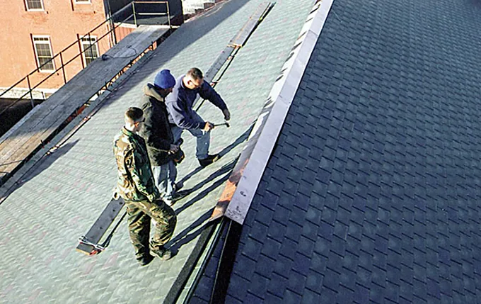 Three roofers installing slate-style shingles and metal ridge trim on a steep roof